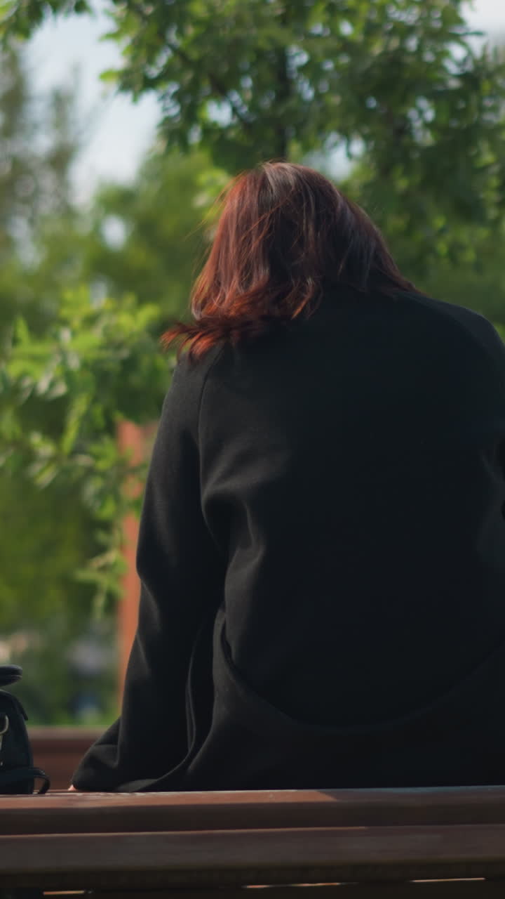 도시 공원의 공원 벤치에 앉아있는 검은 코트와 부츠를 입은 여성의 뒷면, 녹색 나무로 둘러싸여, 놀이터와 함께 흐릿한 배경과 사람들이 돌아다니고 있습니다.