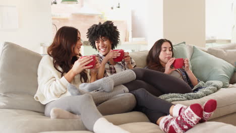 women friends relaxing on cozy sofa, enjoying hot drinks and laughter at home
