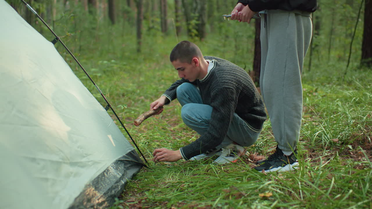 man squats beside tent receiving peg from woman and begins hammering it into forest ground using wood stick while she stands nearby holding remaining pegs during camping setup