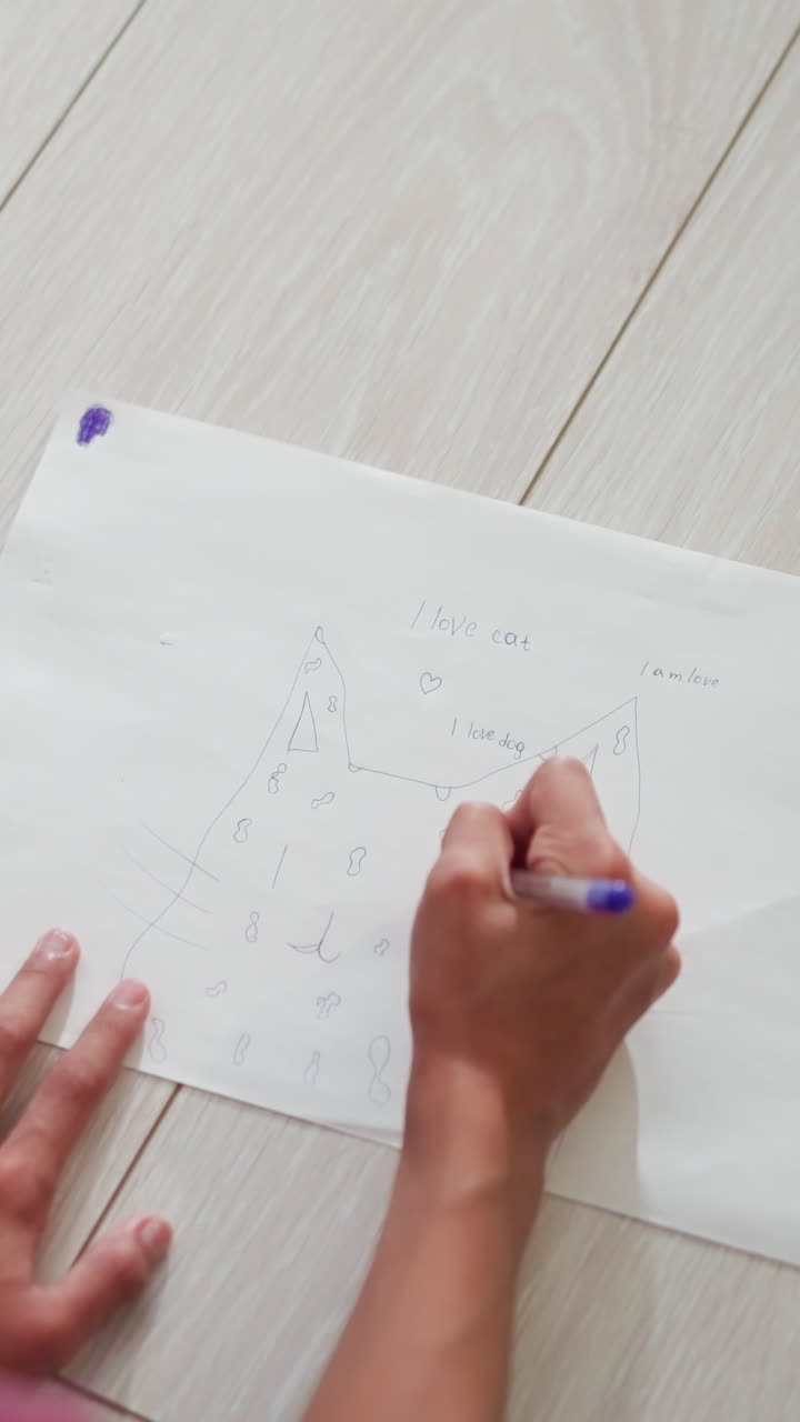 Primer plano de la cabeza de un niño dibujando en el suelo con un bolígrafo azul, con una cuerda roja alrededor de la muñeca, mostrando energía creativa con un dibujo sencillo de una figura de animal y una escritura juguetona que expresa amor por los gatos y los perros.