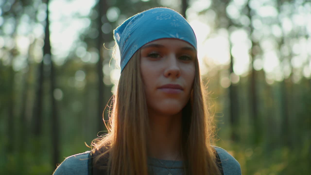 Young woman in blue headscarf tilts her head right with a gentle smile in forest, sunlight casting a warm glow around her face, natural lighting and forest background create serene atmosphere