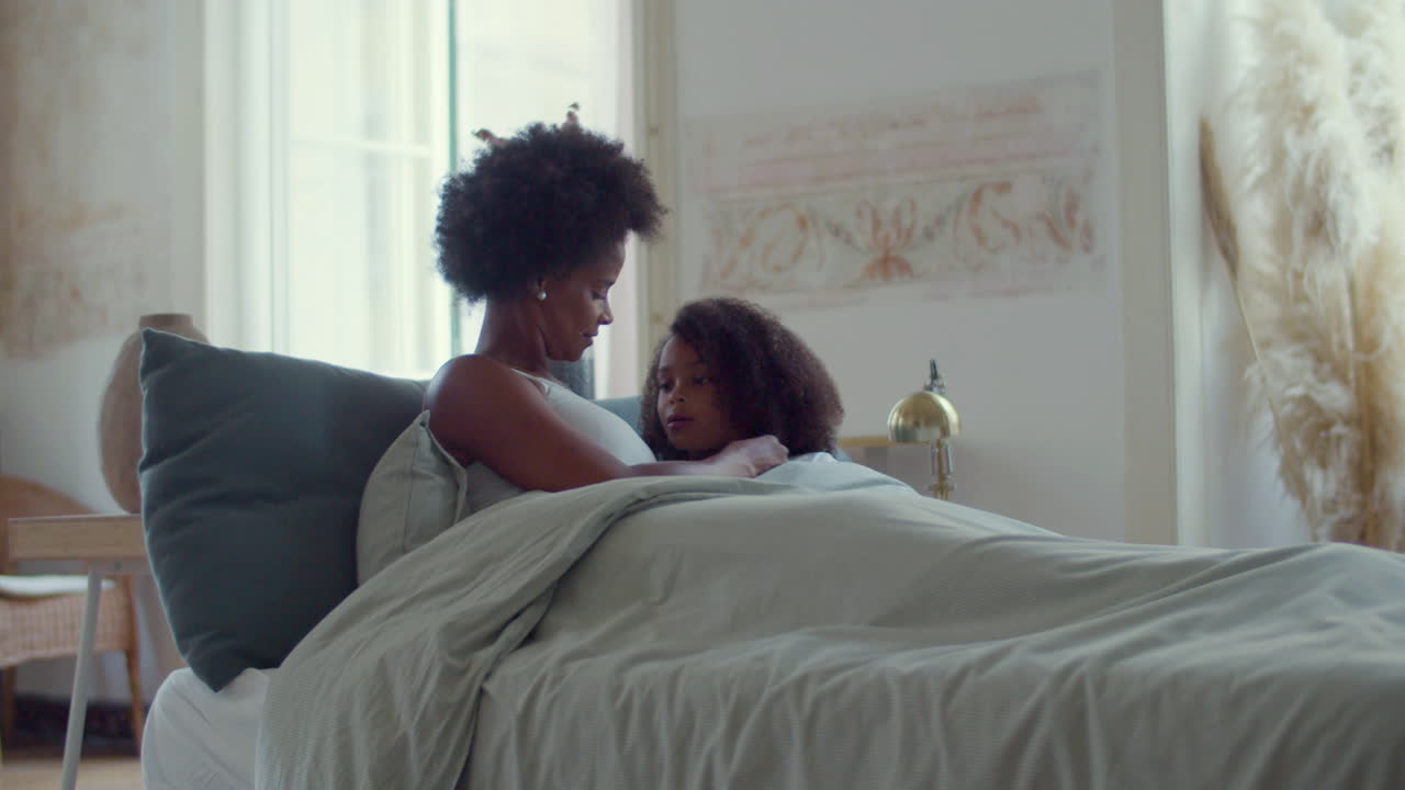 Relaxed African American mum and her little daughter lying in bed on lazy weekend