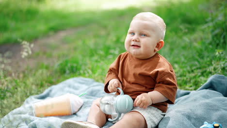 beautiful little boy with blond hair sits on the plaid outdoors. kid looks up holding a water bottle.