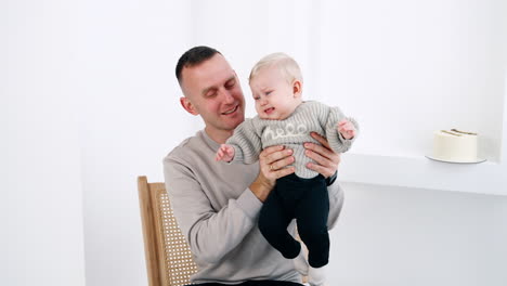father sitting on the chair hold a little infant in hands. kid starts to cry and dad calms him down. white backdrop.