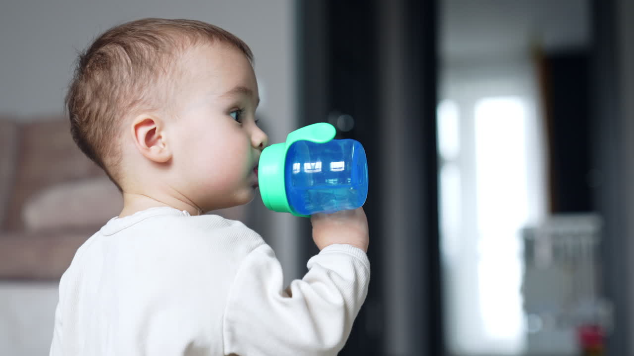 Toddler drinking water from a blue bottle with pacifier. Side view. Baby boy looks up with curiosity. Blurred backdrop.