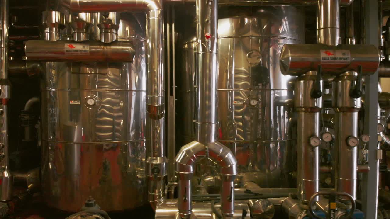 Uma cena ampla dentro de uma cervejaria ou destilaria, mostrando grandes tanques de aço inoxidável, tubulações e válvulas industriais. A câmera se move para a esquerda, revelando as superfícies reflexivas brilhantes e o maquinário complexo.
