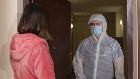 woman opening door at home for doctor in ppe medical biohazard suit, vaccination against coronavirus