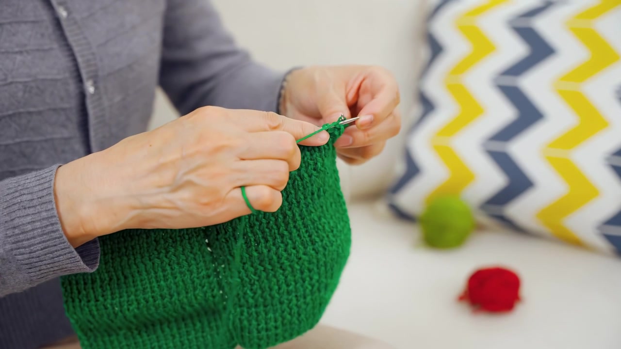 Anziana nonna di talento che lavora a maglia un maglione verde con gomitolo di lana e ferri, con mani rugose, un hobby creativo nel tempo libero, seduta comodamente sul divano. Persone, età e tempo libero.
