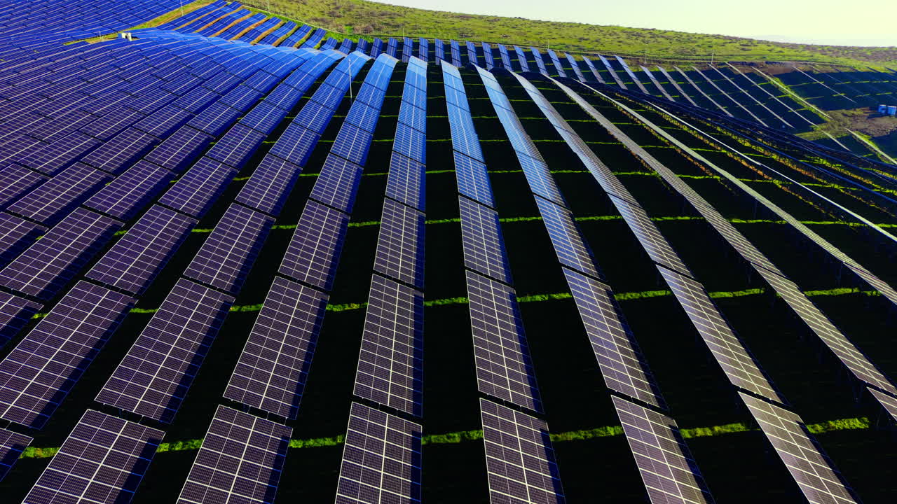 Vista aerea di schiere fotovoltaiche ripetitive che seguono il profilo di verdi colline erbose. Motivi geometrici di file di pannelli solari su colline ondulate.