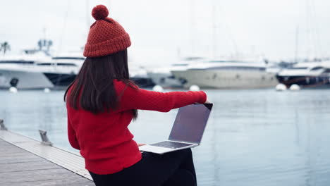 Mujer-Asiática-Sentada-Junto-Al-Agua-En-El-Puerto-De-Valencia,-Abriendo-Su-Trabajo-En-Una-Computadora-Portátil,-Con-Un-Hermoso-Telón-De-Fondo-De-Yates,-Usando-Un-Gorro-Rojo-Que-Complementa-Su-Camisa-Roja-De-Cuello-Alto.