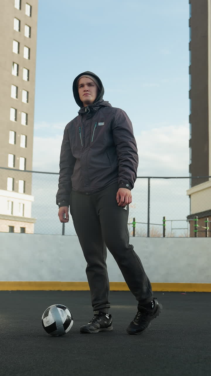 Athlete in hoodie standing confidently in sport arena, turning legs with ball placed in front, urban background features tall modern office buildings under clear sky