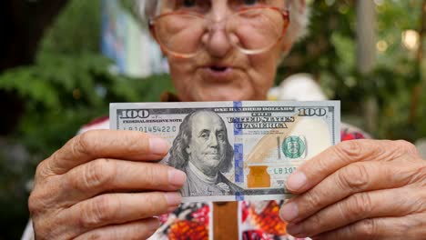 old woman in eyeglasses showing bill of one hundred dollar into camera and smiling outdoor. granny holding cash outside. money concept. close up slow motion