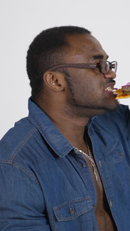 black man in glasses eats pizza. afro american man in blue shirt tastes a piece of delicious pizza on white studio background. profile view. vertical video