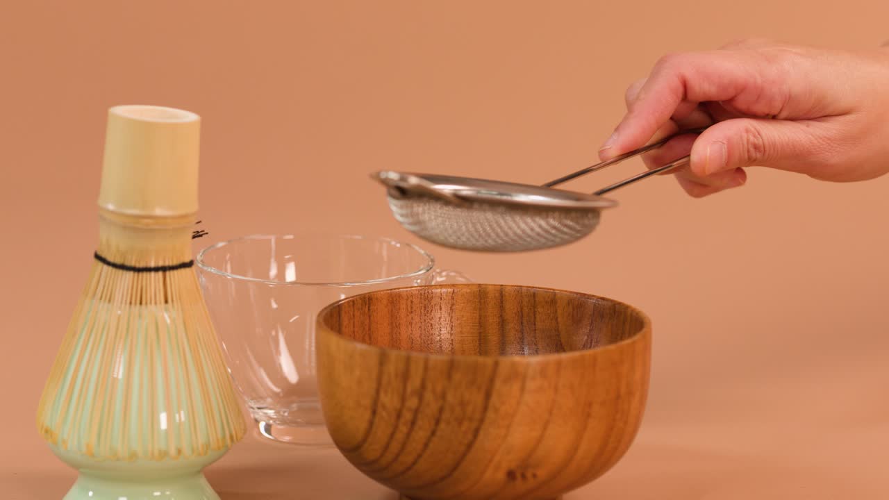 Una mano setaccia la polvere di tè verde con un colino in una ciotola di legno, illuminazione delicata da studio, camera statica