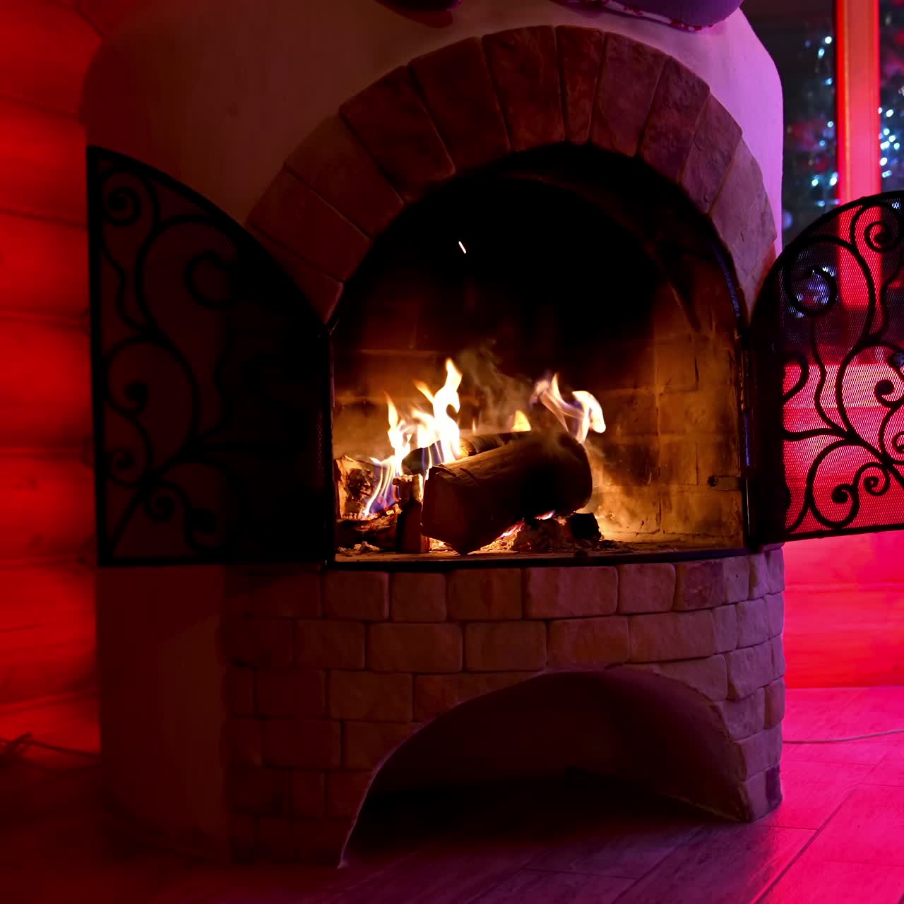 Fireplace of a modern design next to the Christmas tree. Burning firewood in mantel in the living room at Christmas. Coziness and relaxation in winter holidays. Red lighting