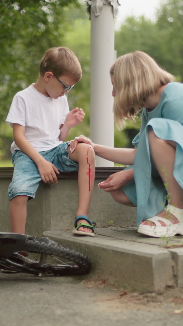 una madre preocupada se arrodilla al lado de su hijo herido, limpiando una herida sangrienta en la rodilla con un pañuelo, mientras el niño persigue moscas de la herida