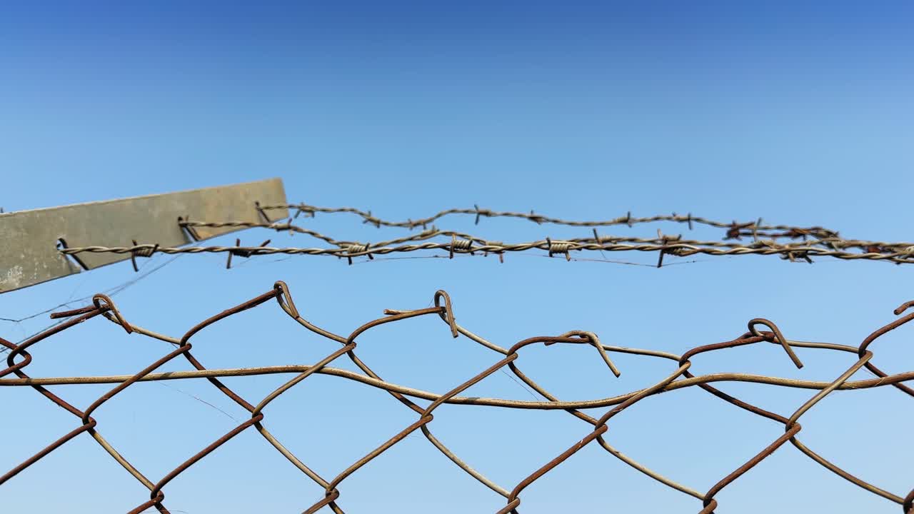 Tracking shot of a rusted chain-link fence topped with barbed wire against a vivid blue sky, revealing weathered textures and a sense of isolation