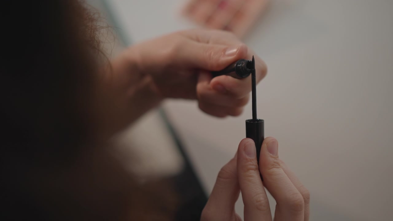 Tight close-up shows hands lifting mascara wand from open tube with visible upward motion preparing for eyelash application; shallow depth of field blurs background and softens facial area while