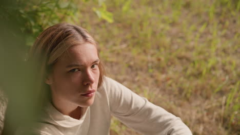 young lady seated on ground under tree with hand resting on knee, looking up thoughtfully with gentle expression, soft background of swaying leaves and natural surroundings