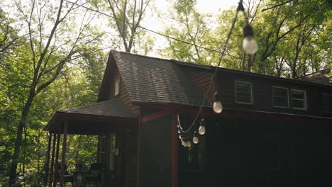 panning view of a maroon cabin surrounded by woods, adorned with string lights in summer