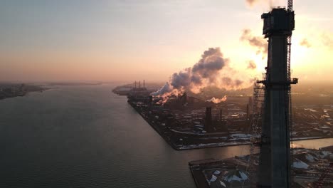 New-Gordie-Howe-bridge-tower-under-construction-with-smoking-plant-behind-during-susnet,-aerial-drone-view