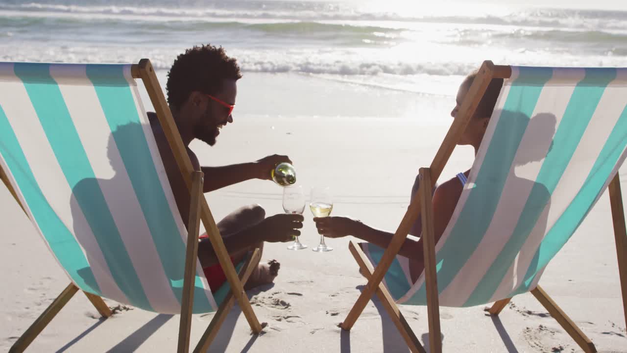 African american couple drinking wine together sitting on deck chairs at the beach