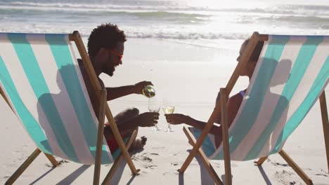 african american couple drinking wine together sitting on deck chairs at the beach