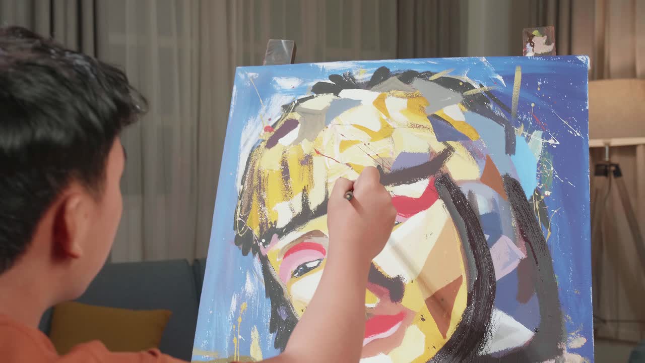 Hind View Of An Asian Artist Boy In Wheelchair Holding Paintbrush And Mixing It With Colour Before Painting A Girl'S Face On The Canvas