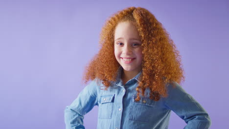 studio portrait of smiling girl with red hair standing against purple background with hands on hips