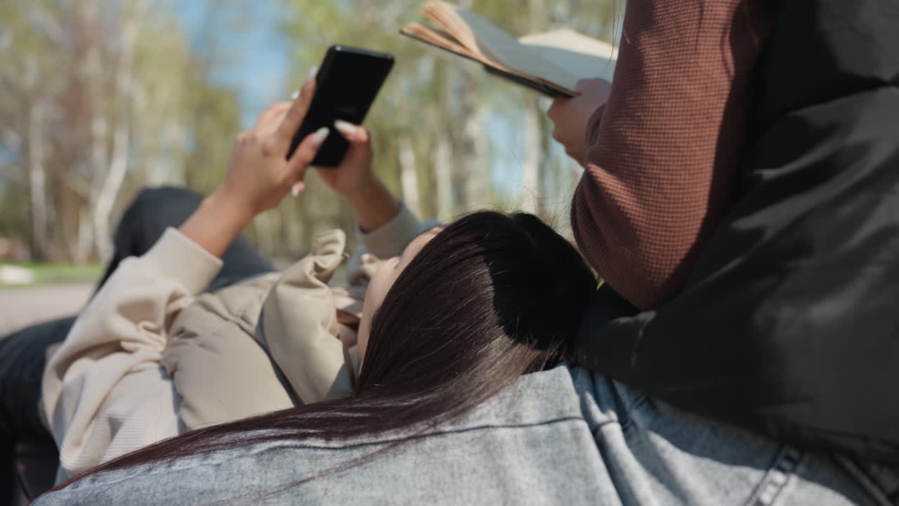 여자가 벤치에 앉아 책을 읽고 있고, 친구는 스마트폰을 보고 있네요. 자작나무와 햇살이 비치는 공원 산책로, 캐주얼한 청바지와 스웨터를 입고 편안한 분위기로 야외에서 만나 우정을 나누는 모습이 자연스럽게 담겨 있어요.