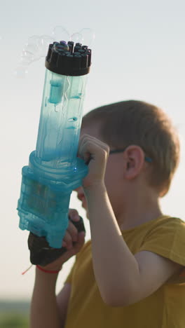 un niño joven con una camisa amarilla y gafas se encuentra al aire libre, sosteniendo alegremente una pistola de burbujas de plástico azul que libera burbujas en el aire. en un campo abierto y brillante con cielos despejados