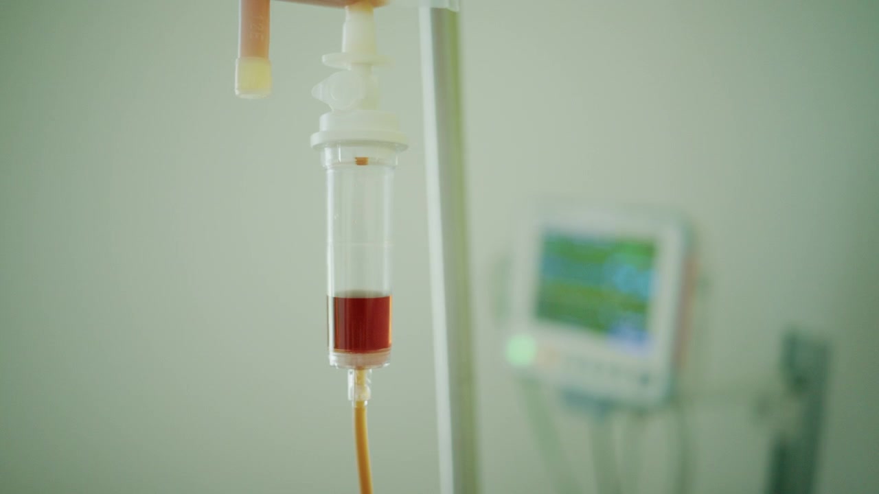 Blodtransfusionspose og IV-drop, der leverer behandling i et hospitalsmiljø, med en patientmonitor i baggrunden
