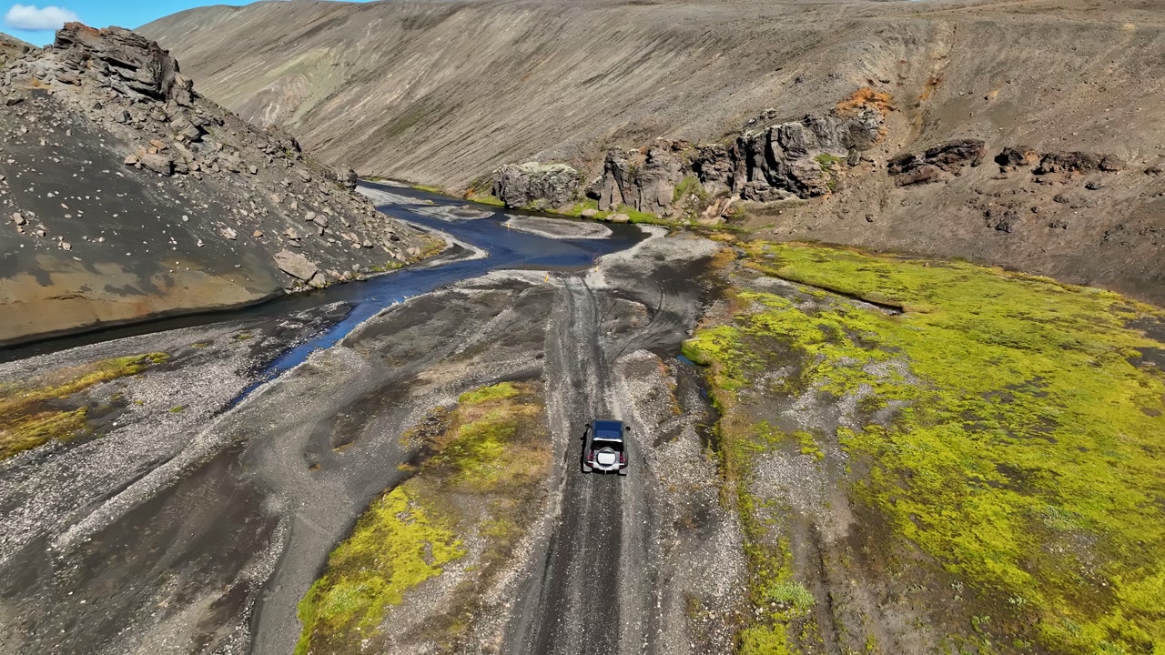 Aerial view of an SUV driving through an Icelandic volcanic valley with winding river channels, dark gravel tracks, and vivid green moss, capturing remote overland adventure in the highlands.