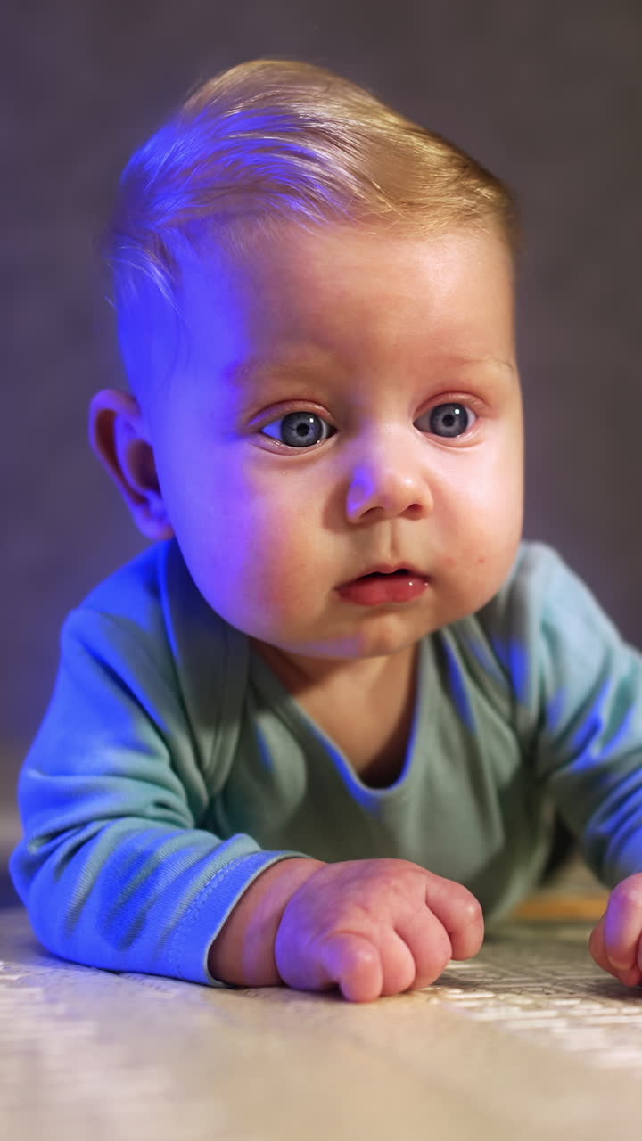 Lovely big-eyed baby looking ahead with surprise. Beautiful infant lies on the floor close up. Vertical video