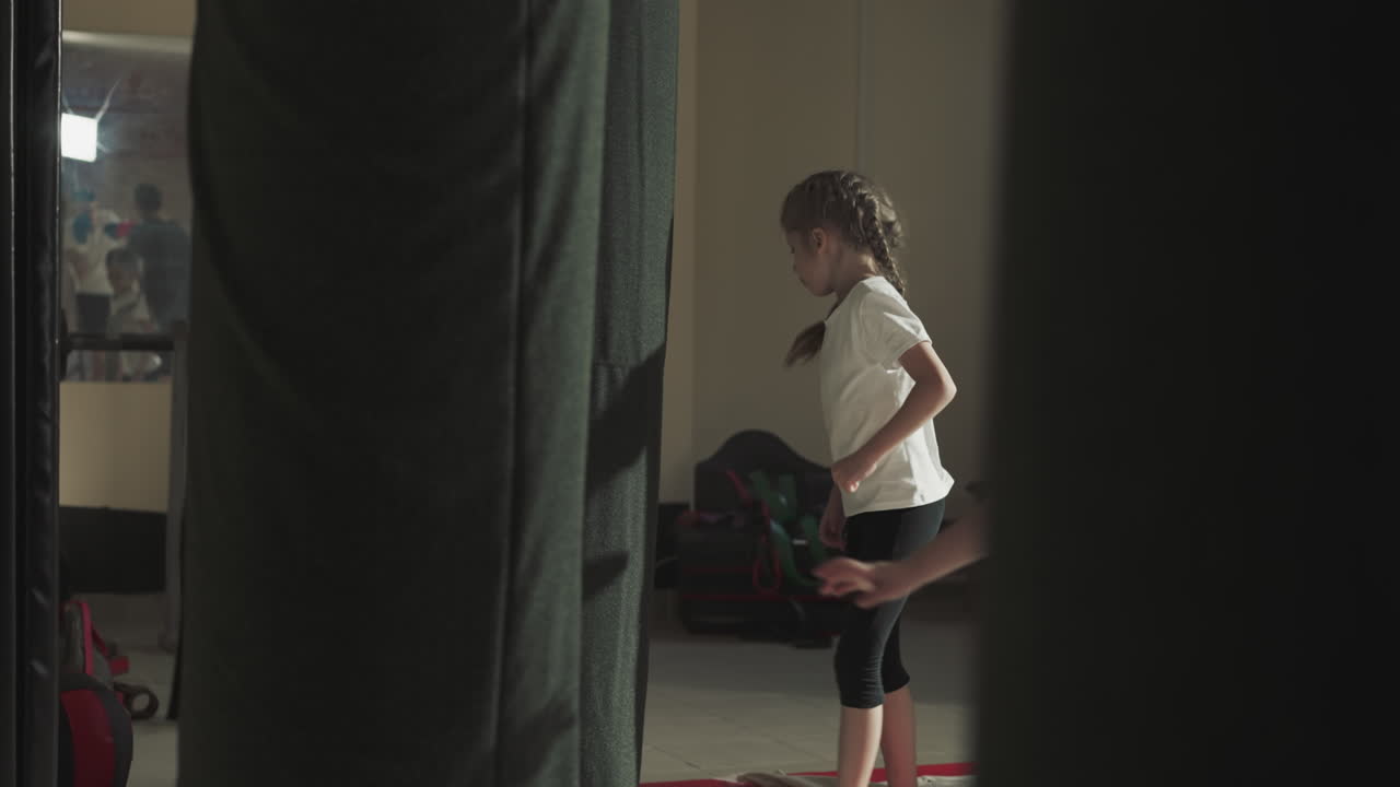 Kid kicks punching bag jumping with teammates in gym. Active little girl exercises with equipment during group lesson in sports club slow motion