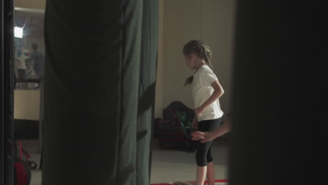 kid kicks punching bag jumping with teammates in gym. active little girl exercises with equipment during group lesson in sports club slow motion