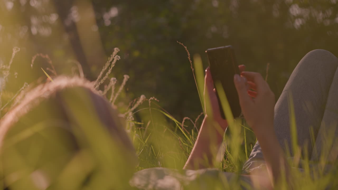 Close-up of young lady lying in grassy field, holding and looking at her phone, warm sunlight filters through surrounding grass, creating a glowing, peaceful atmosphere