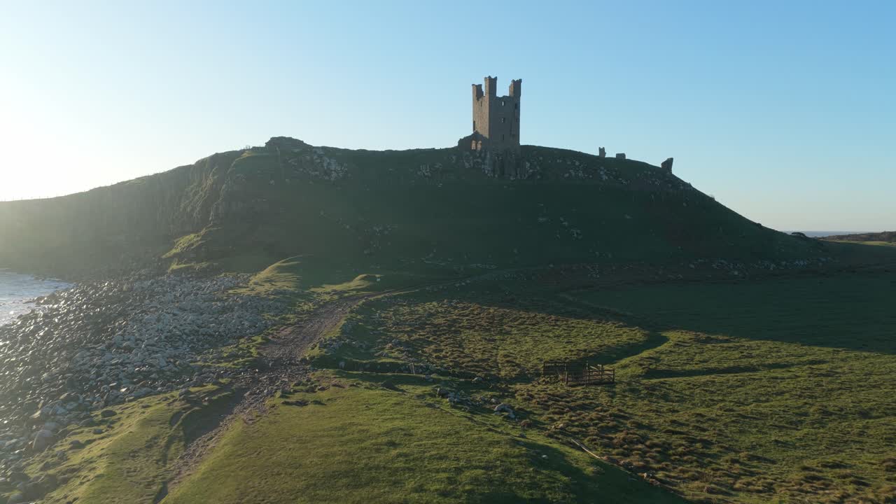 Baglæns luftoptagelse af Dunstanburgh Slot ved solopgang.