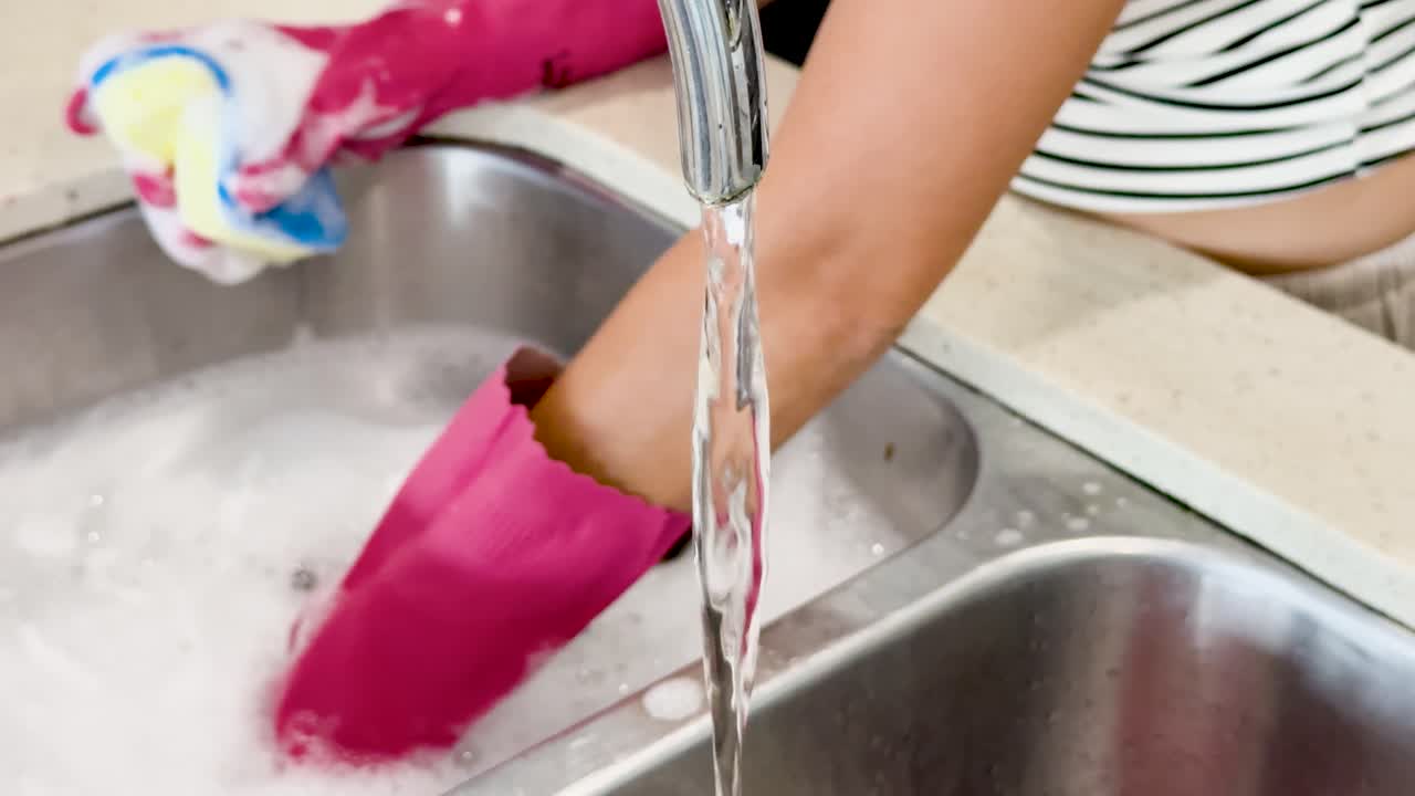 A person wearing pink rubber gloves scrubs and rinses a ceramic mug under running water in a bright kitchen, using a colorful sponge