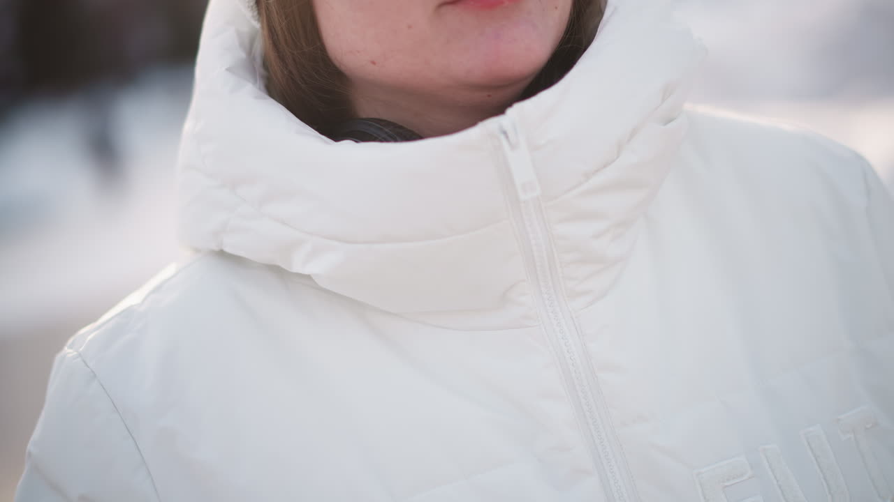 Young woman in winter coat wearing headphones zips up puffer jacket at cold outdoor setting, hands bringing zipper to chin, crisp light reflecting on jacket and skin, ready for chilly adventure