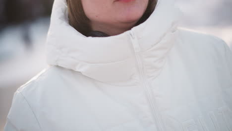young woman in winter coat wearing headphones zips up puffer jacket at cold outdoor setting, hands bringing zipper to chin, crisp light reflecting on jacket and skin, ready for chilly adventure