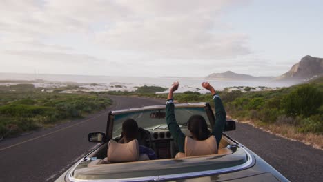 Vista-Trasera-De-Una-Pareja-Diversa-Conduciendo-En-Un-Día-Soleado-En-Un-Auto-Convertible-Levantando-Los-Brazos-En-El-Aire