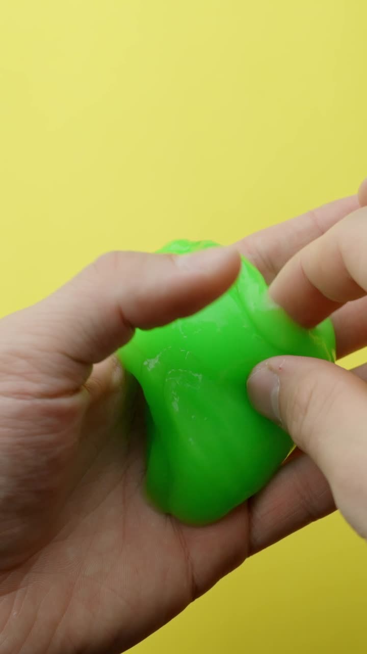 Close-up of hands squeezing and kneading a bright green slime against a plain yellow background. Vertical