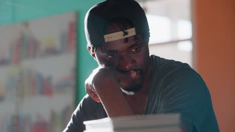 Thoughtful young man resting chin on hand, gazing downward in deep contemplation near blurred stack of books with sunlight streaming through bright window in background