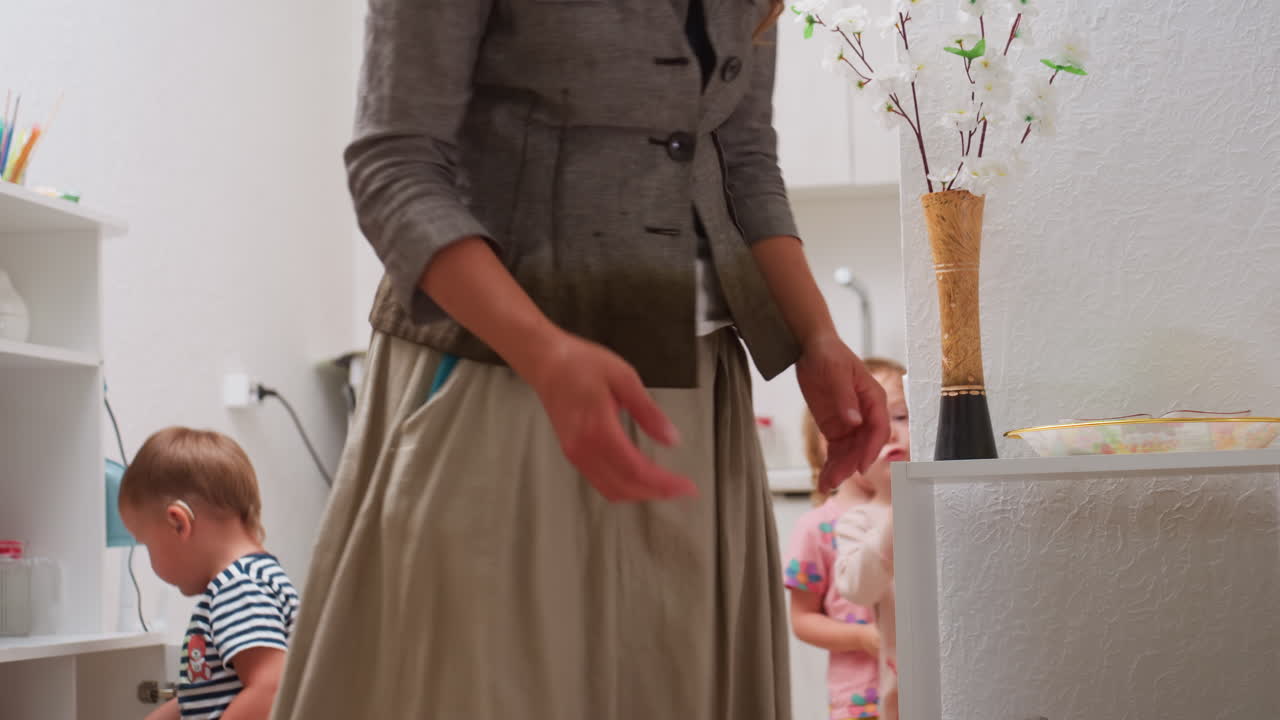 Kindergarten children moving around kitchen as teacher organizes items on shelf, showing interaction, supervision, and daily preschool routine with warm indoor setting of early learning environment