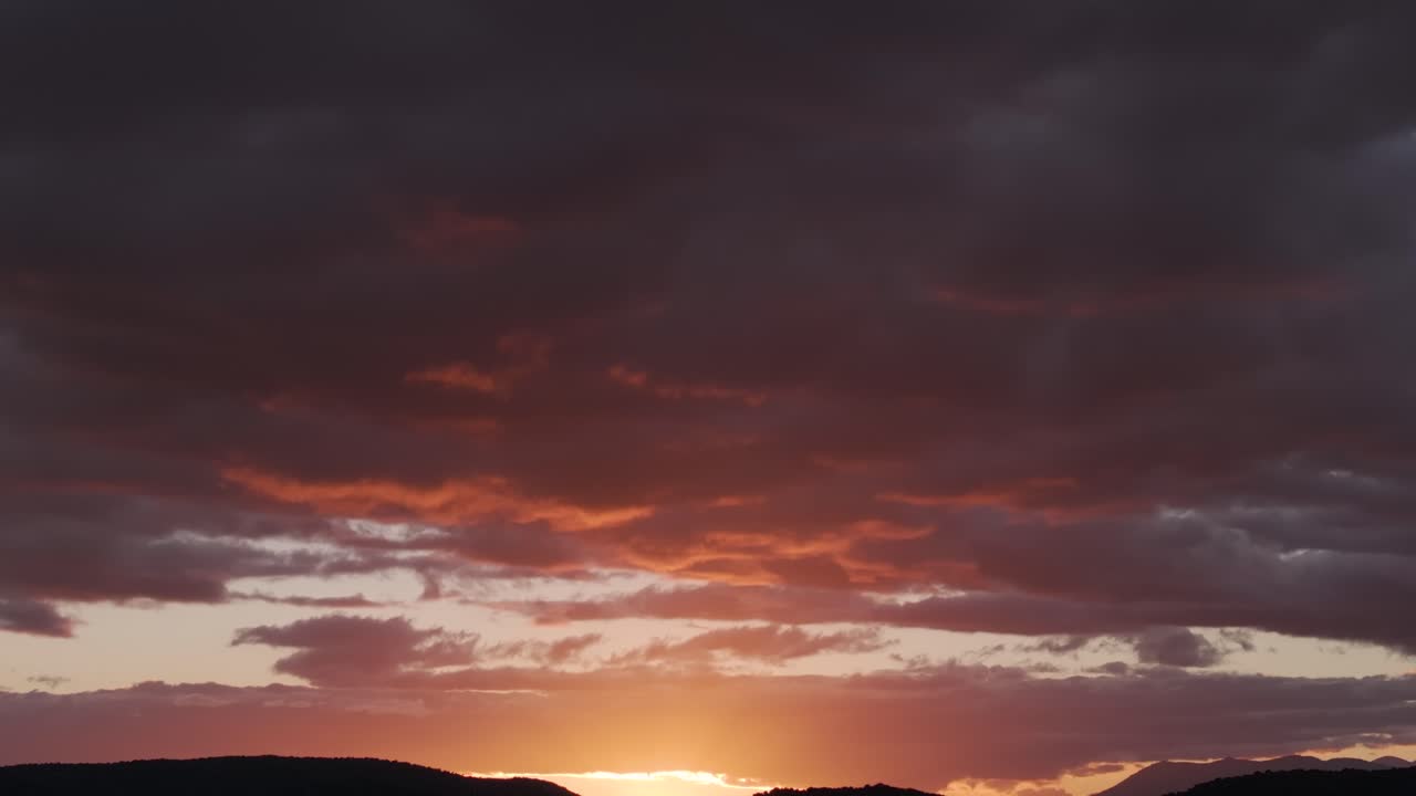 Descending drone flight through dark bluish-gray clouds into vivid orange sunset light, ending over deep mountain silhouettes against the glowing horizon.