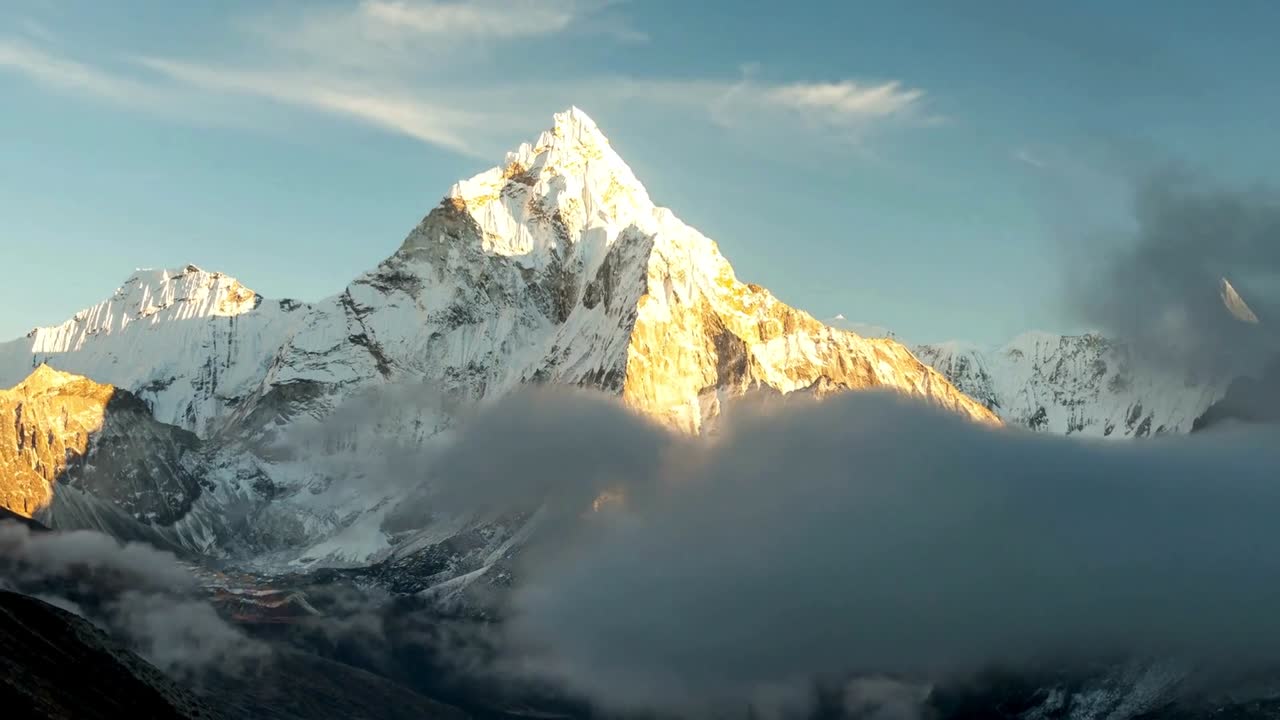 A stunning time-lapse of a majestic snow-covered mountain peak bathed in golden sunrise light. Soft clouds move gracefully at the base, highlighting the grandeur and beauty of the rugged alpine