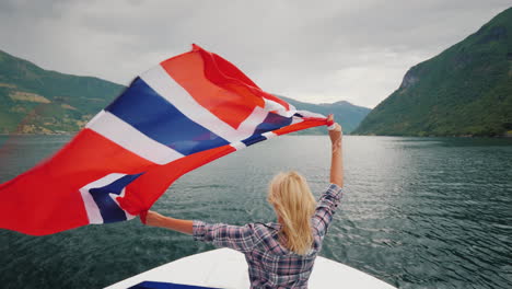 eine aktive frau mit einer großen norwegischen flagge steht am bug eines kreuzfahrtschiffes bei einer fjordkreuzfahrt