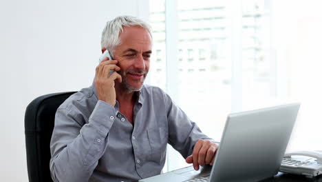 Casual-businessman-working-on-laptop-and-talking-on-phone-at-desk