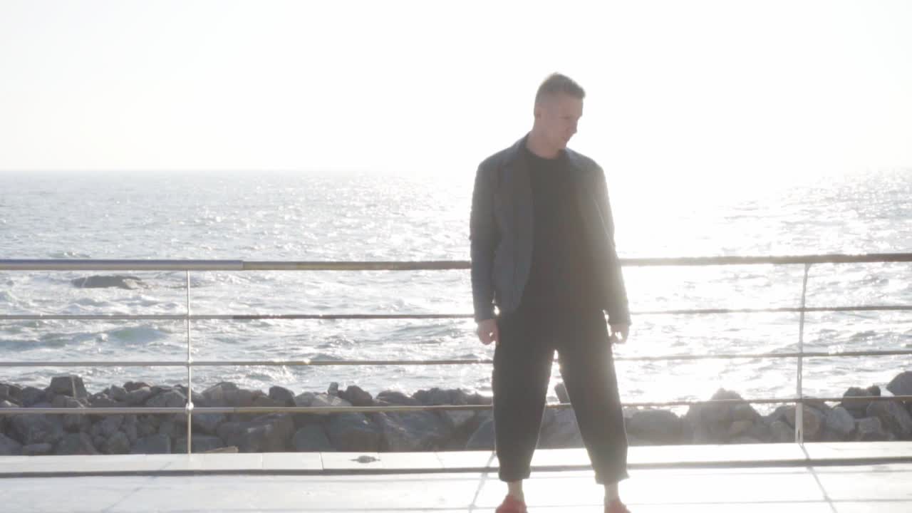 가죽 재 ⁇ 을 입은 멋진 젊은이가 바 ⁇ 가의 롱보드에서 스케이팅을 하고 있습니다.